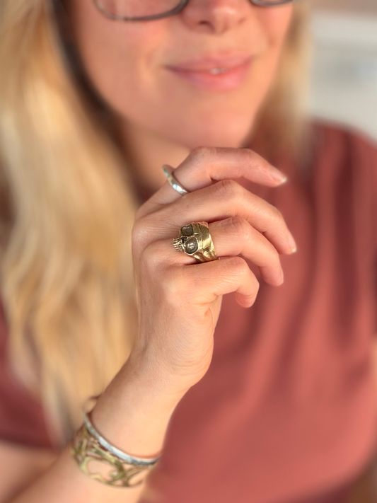 A close-up lifestyle shot of a woman wearing a chunky golden brass ring with a central skull detail on her finger, paired with a patterned bracelet and a simple silver band on another finger.