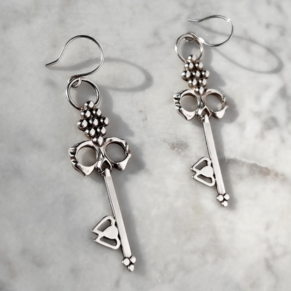 Sterling Silver Skull Key Earrings that dangle from handmade French hooks lay on white marble