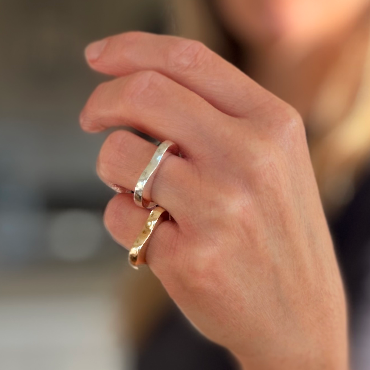 A close-up of a hand wearing two rings: a smooth silver band and a smooth brass band set against a softly blurred background.