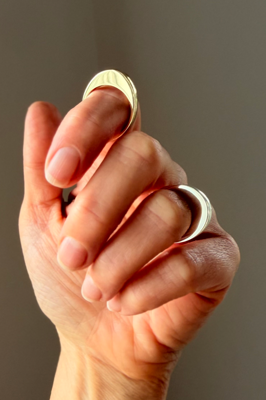 A close-up of a hand wearing two minimalist curved rings, one in polished brass and the other in polished silver, against a neutral gray background.