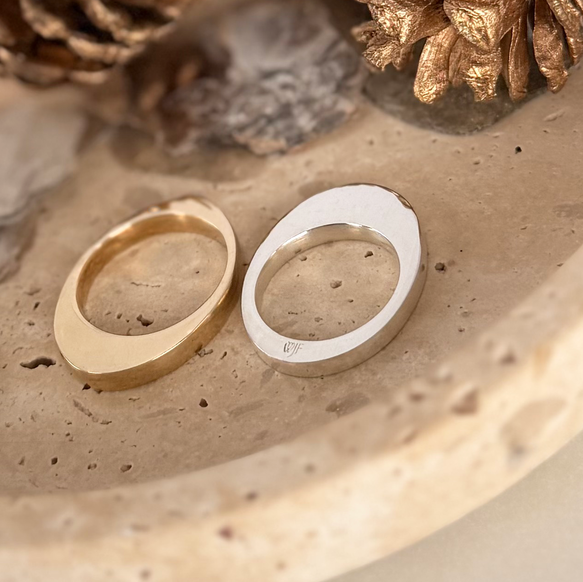 Two polished egg-shaped rings nested together, one in gold and the other in silver, displayed on a textured beige stone surface with decorative pinecones.