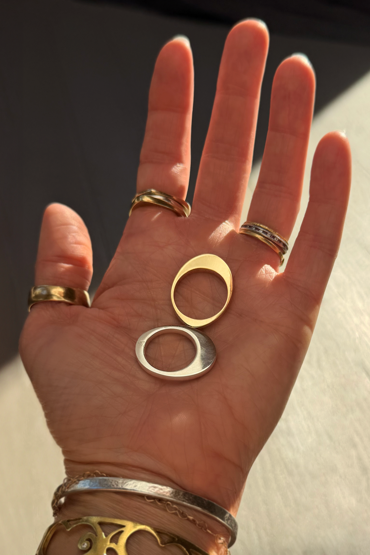A close-up of a hand holding two shining egg-shaped bands in its palm, with light and dark shadows in the background. The hand is adorned with multiple gold and silver rings, including gold and silver bracelets.