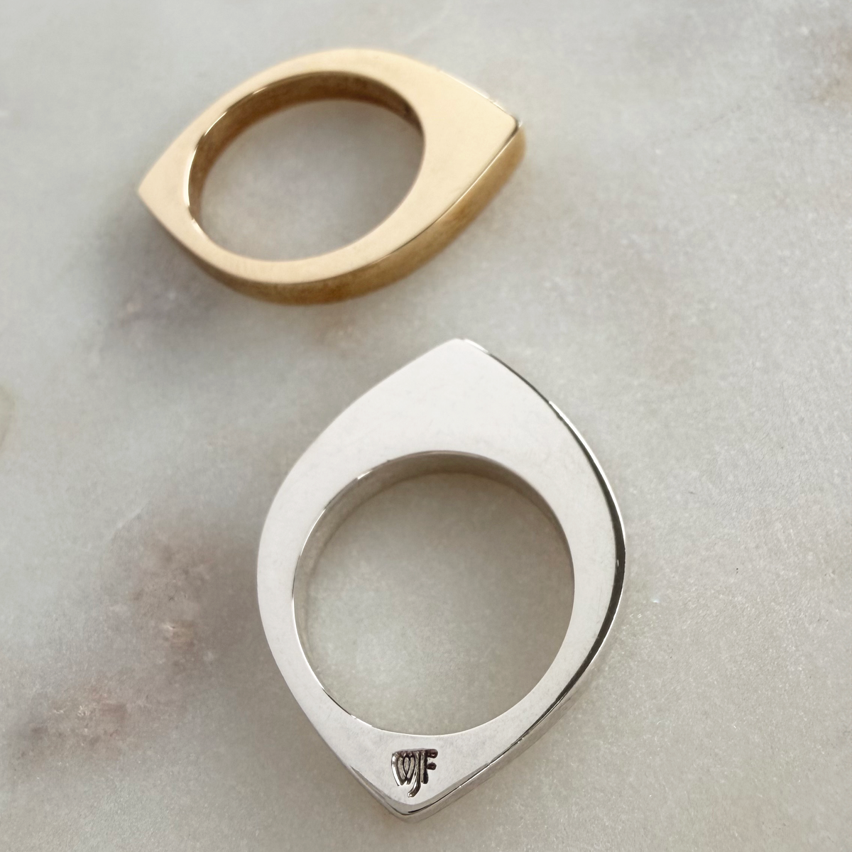 Two modern eye-shaped rings, one in polished gold and one in polished silver, displayed on a light gray marble surface. The silver ring features a small engraved maker's mark at the bottom.