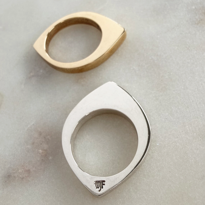 Two modern eye-shaped rings, one in polished gold and one in polished silver, displayed on a light gray marble surface. The silver ring features a small engraved maker's mark at the bottom.