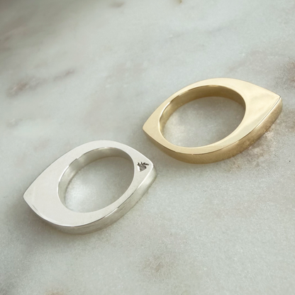Two modern eye-shaped rings in polished silver and gold, displayed on a light gray marble surface.