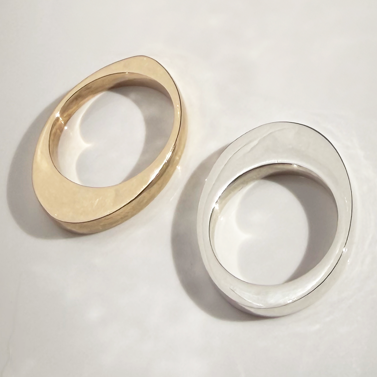 Two minimalist oval ring bands, one in polished yellow gold and one in polished silver, displayed on a light gray surface with soft shadows.