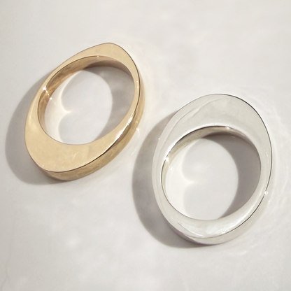 Two minimalist oval ring bands, one in polished yellow gold and one in polished silver, displayed on a light gray surface with soft shadows.