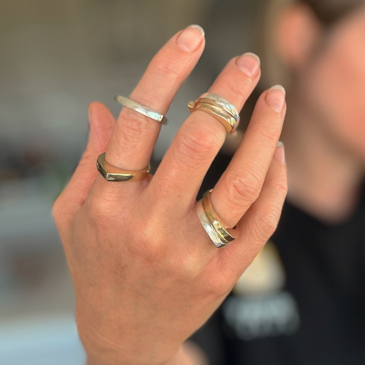 A close-up of a hand wearing multiple minimalist metal rings, including silver and gold bands, with a softly blurred person in the background.