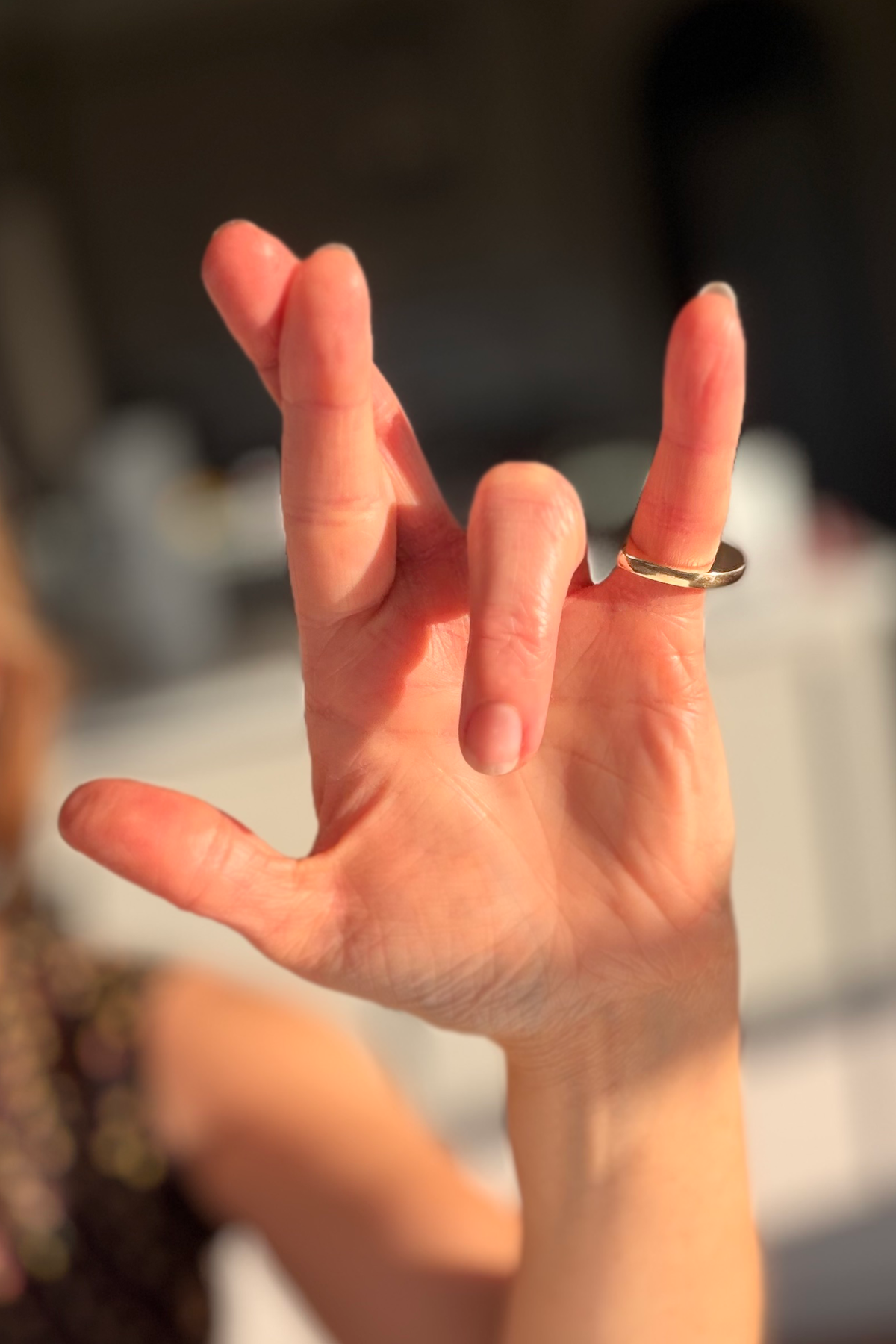 A close-up of a person's hand signing "I really love you" in ASL, wearing a simple gold band ring on one finger, with warm sunlight casting shadows across the palm.