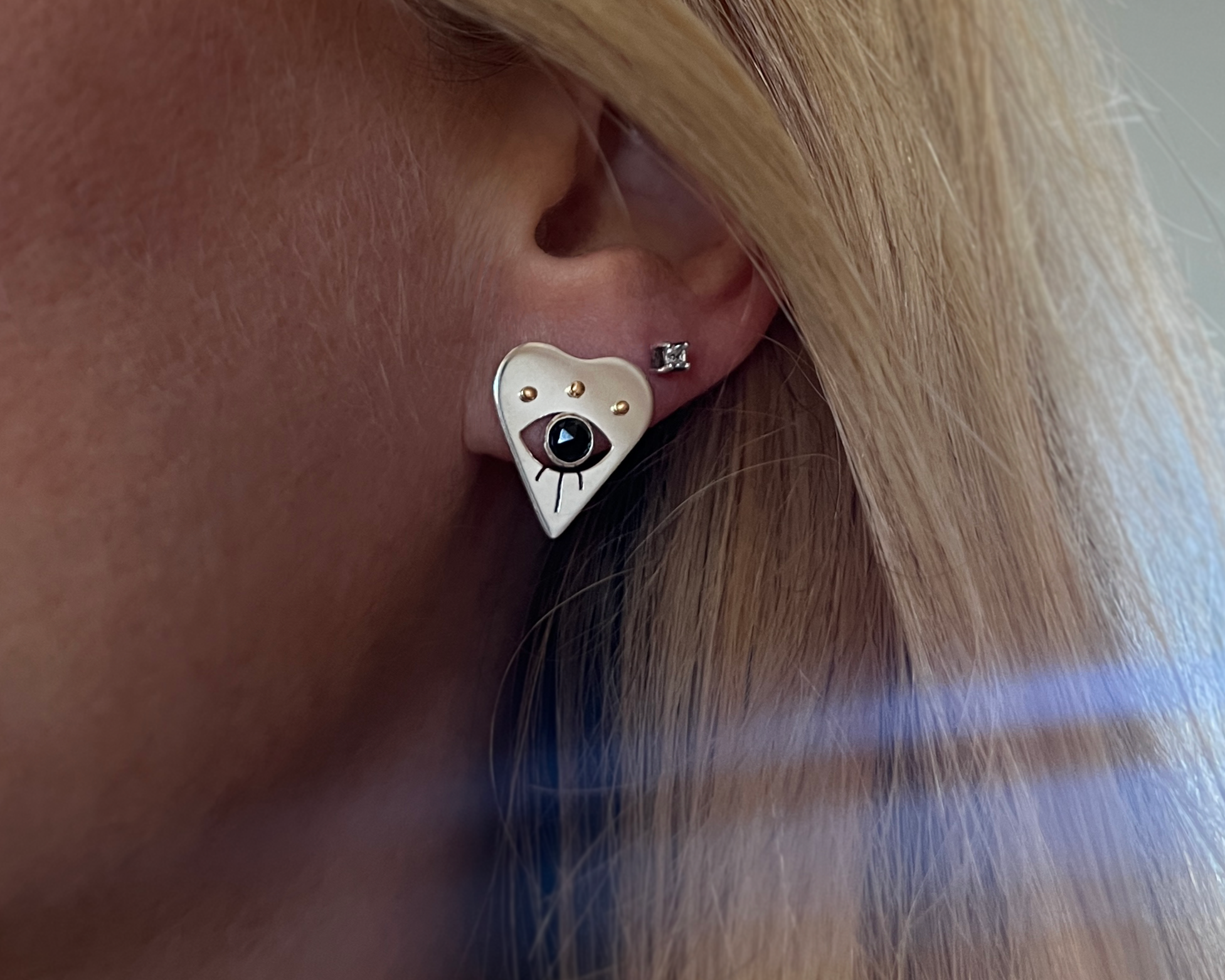 Heart-shaped evil eye earring with a black spinel worn by a person with blonde hair.