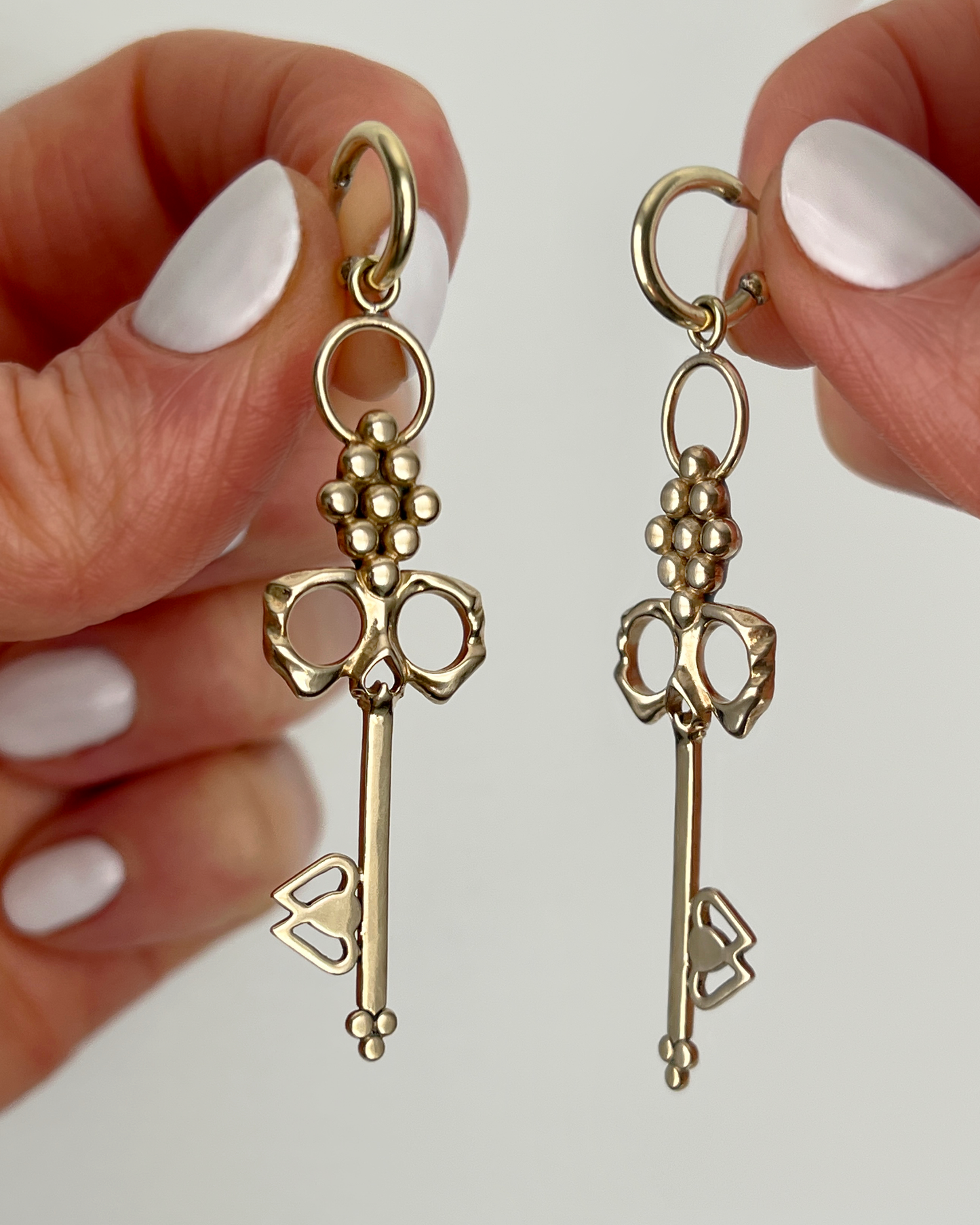 Two hands holding up Brass Skull Key Earrings that dangle from small detachable Huggie Hoops 