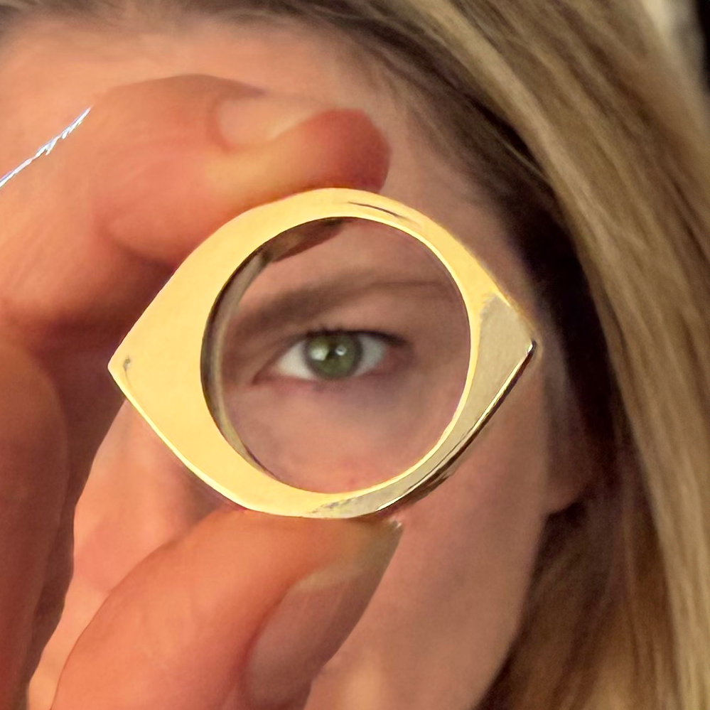A close-up lifestyle shot of a person holding a gold ring up to their eye, demonstrating its eye shape. The ring has a polished, reflective gold finish and a unique eye-shaped silhouette.