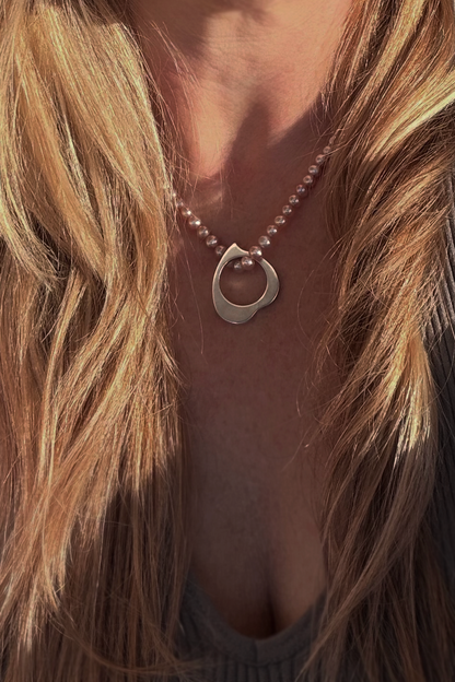 A close-up of a person wearing a delicate heart-shaped, polished silver pendant on a strand of small pinkish pearls, framed by long blonde hair.