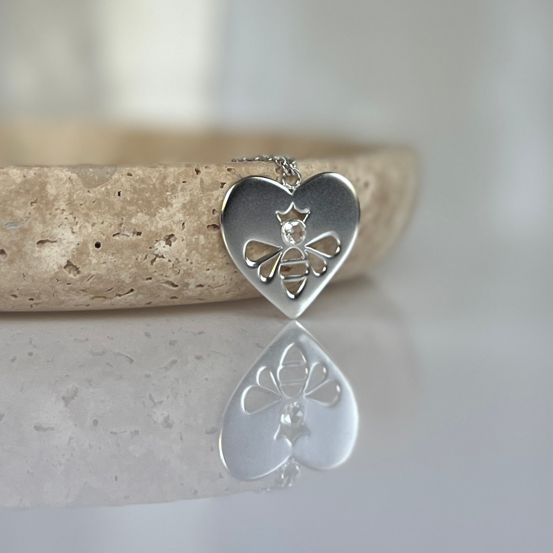 Queen Honey Bee cutout in sterling silver heart medallion necklace with round rose cut white topaz draped over jewelry dish on reflective surface