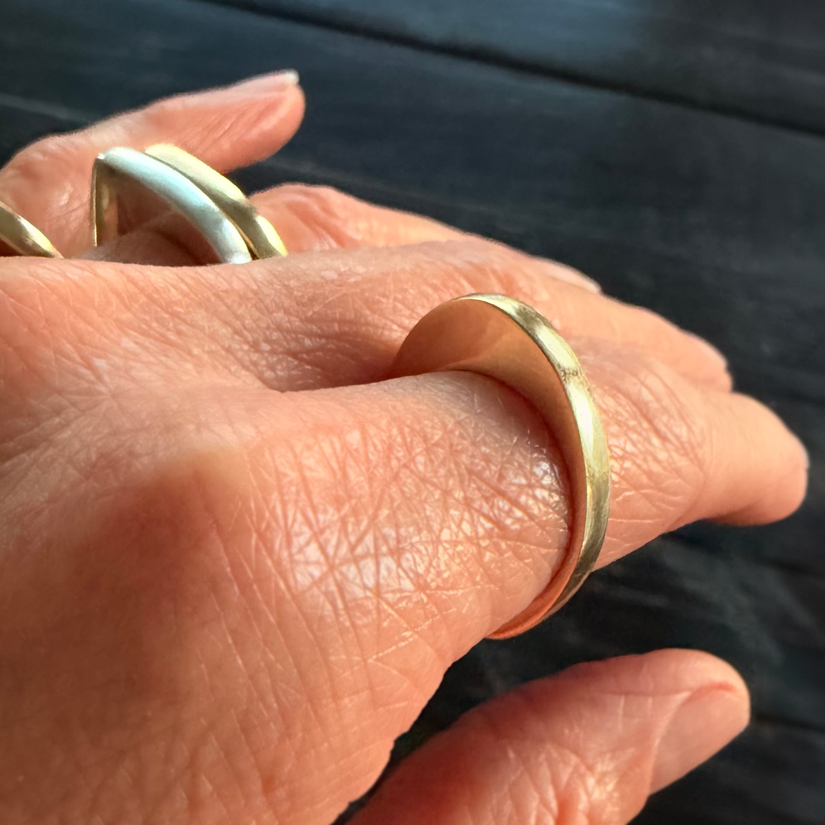 A close-up of a hand wearing a simple, polished gold band ring, with other rings partially visible on the fingers, set against a dark textured wooden background.