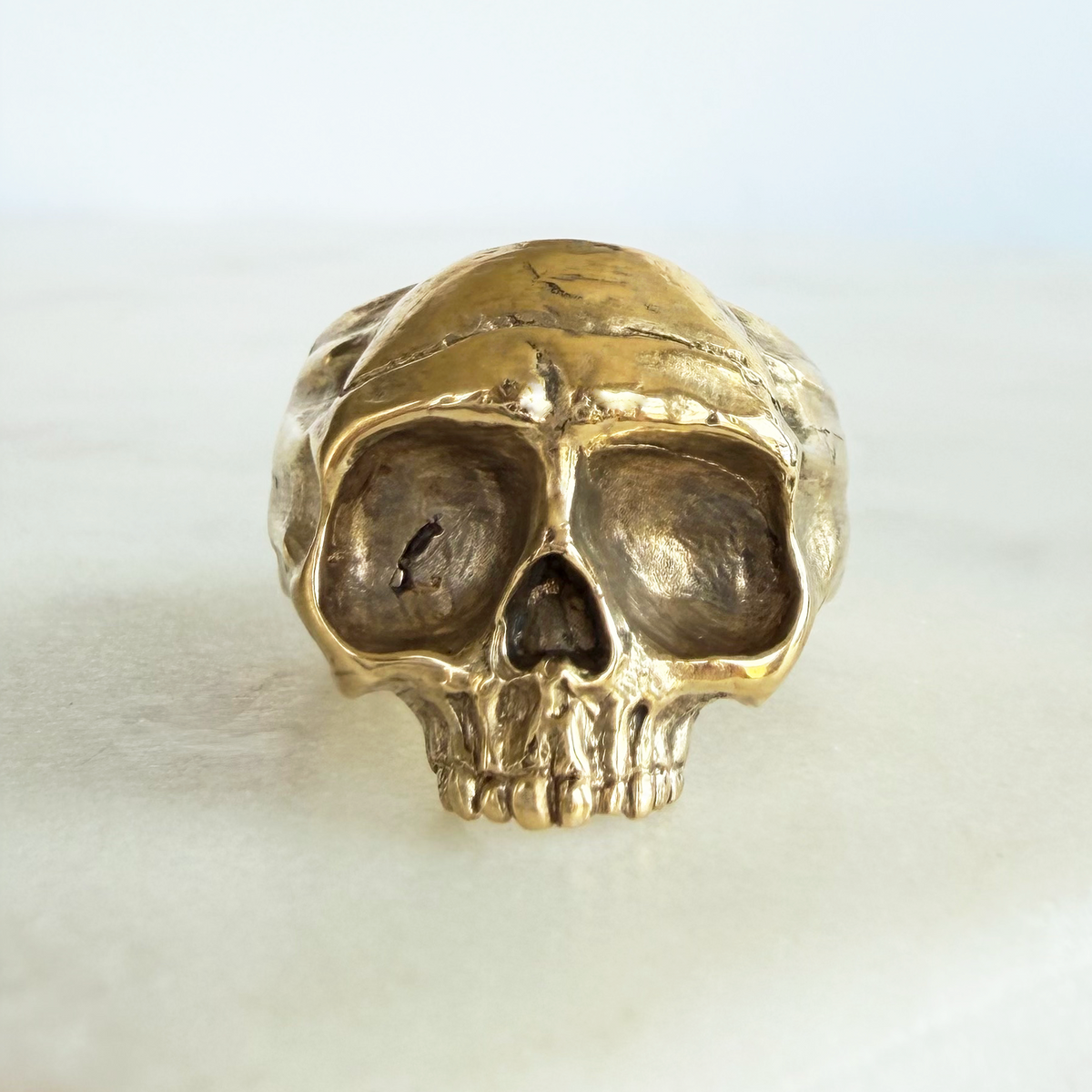 A textured golden brass skull ring displayed on a light marble surface against a white background.