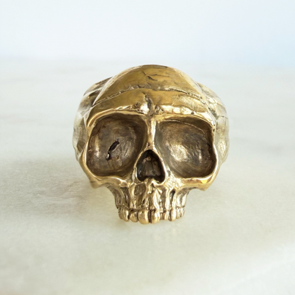 A textured golden brass skull ring displayed on a light marble surface against a white background.