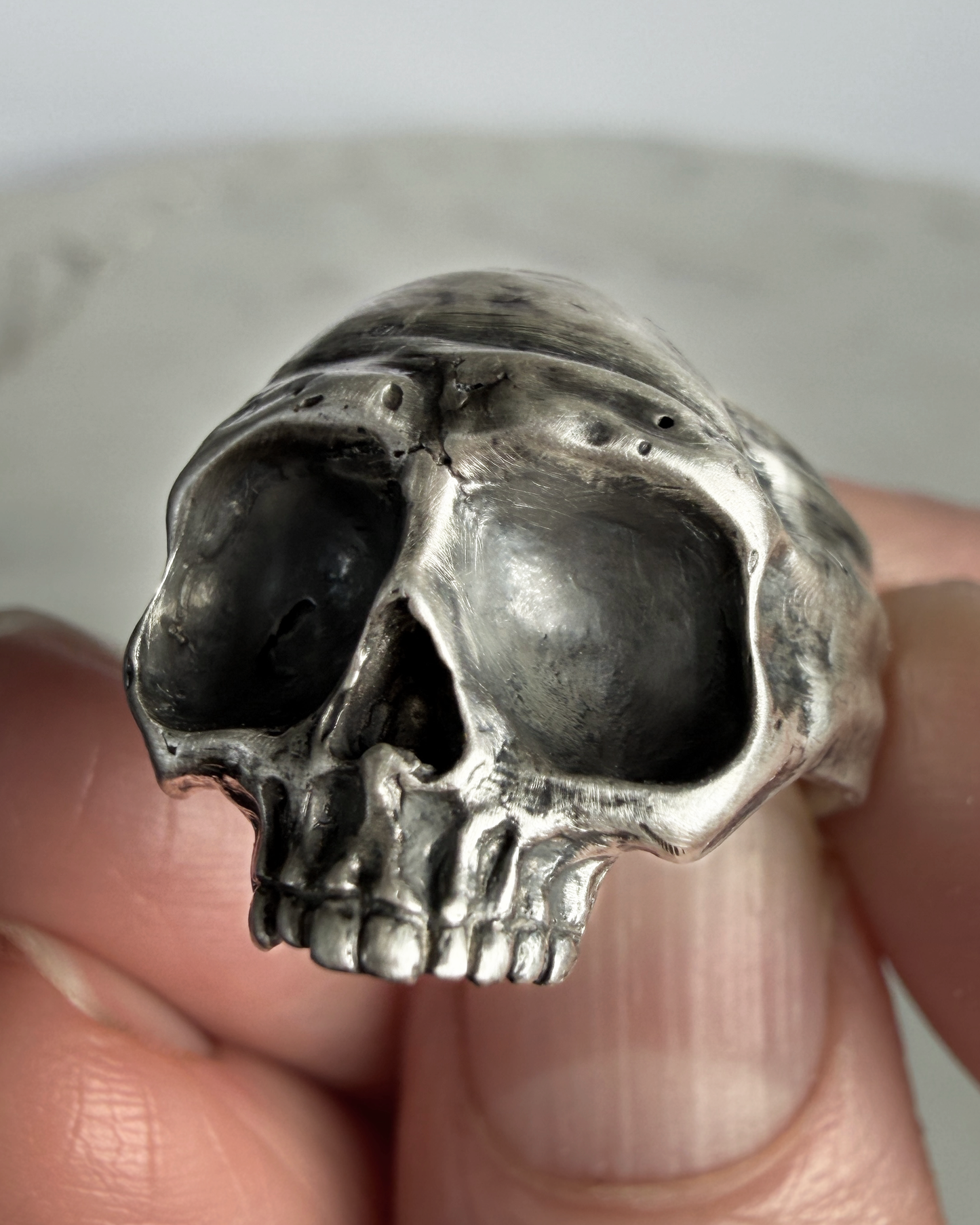 Silver skull-shaped ring held between fingers against a neutral background