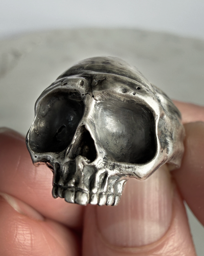 Silver skull-shaped ring held between fingers against a neutral background