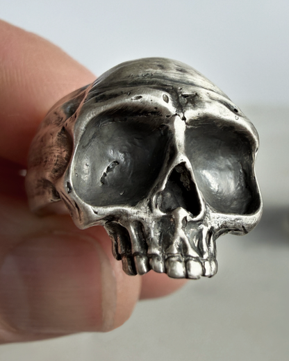 Silver skull-shaped ring held between fingers against a neutral background
