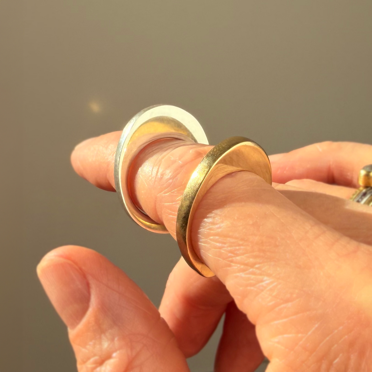 A close-up of a hand wearing two minimalist stacked rings: a shiny gold band and a polished silver band reflecting light against a soft, warm gray background.