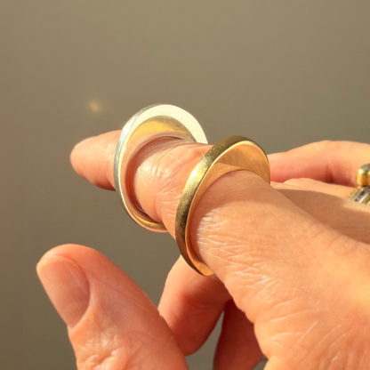 A close-up of a hand wearing two minimalist stacked rings: a shiny gold band and a polished silver band reflecting light against a soft, warm gray background.