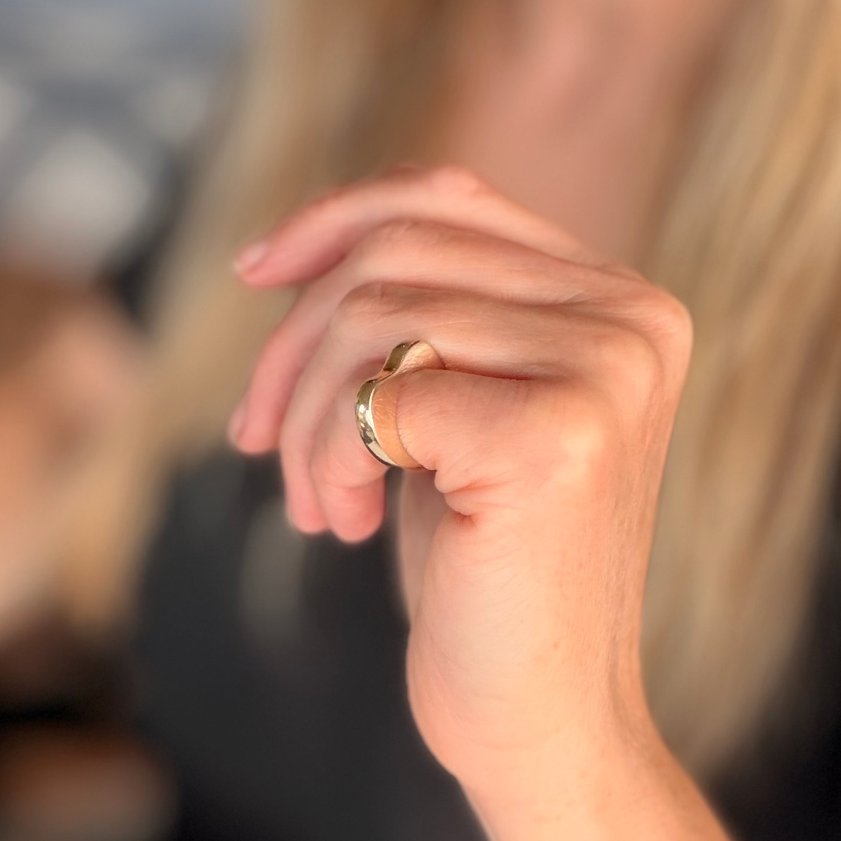 A close-up of a person's hand wearing a simple gold band ring, with soft-focus blonde hair and a dark top in the background.
