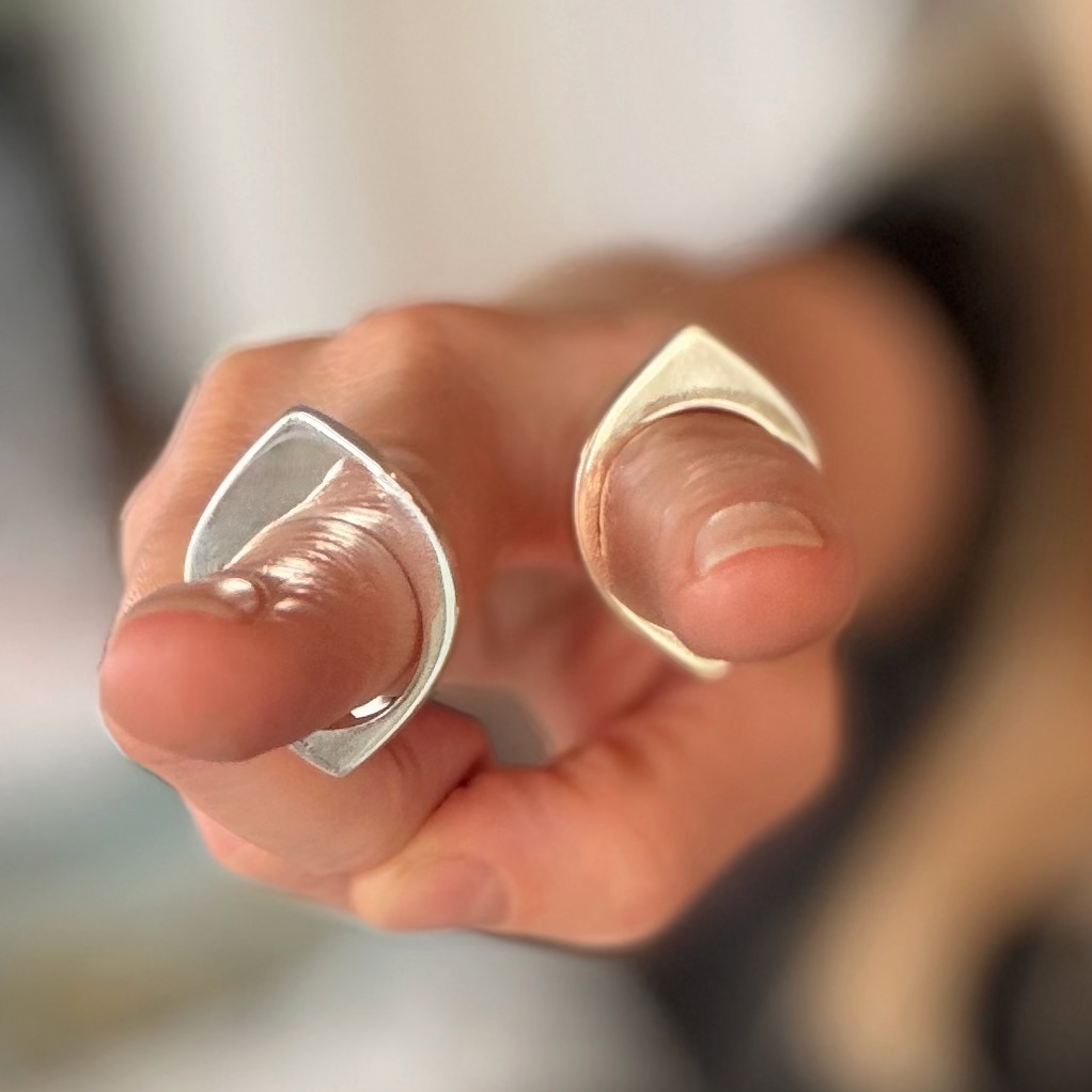 A close-up of a hand wearing two eye-shaped rings on the fingers, one in silver and one in gold, with a soft blurred background.