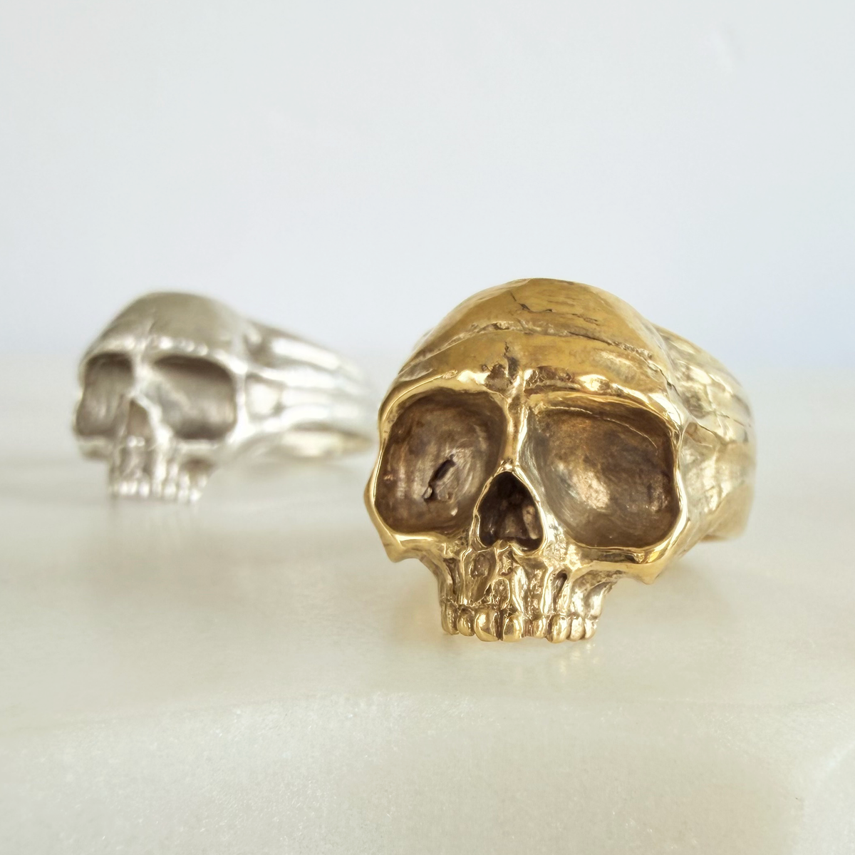 Two skull-shaped rings, one in polished golden brass and one in silver, displayed on a light cream surface with a soft white background.