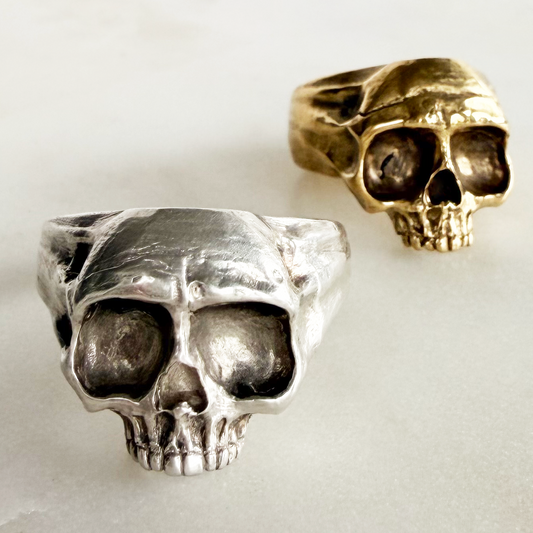 Two skull-shaped rings, one in silver and one in gold tone, displayed on a light gray marble surface.
