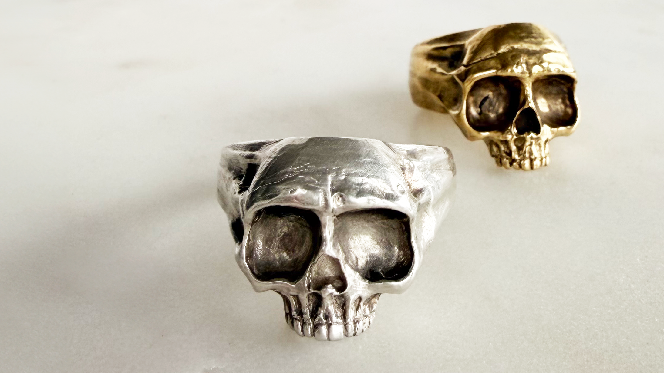 Two skull-shaped rings, one in silver and one in golden brass, displayed on a light gray marble surface.