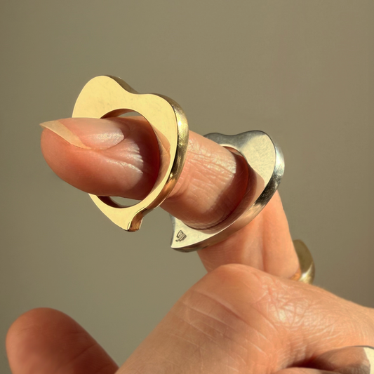 Two heart-shaped rings up-close on a finger, one in gold and one in silver with a blurred, neutral background.
